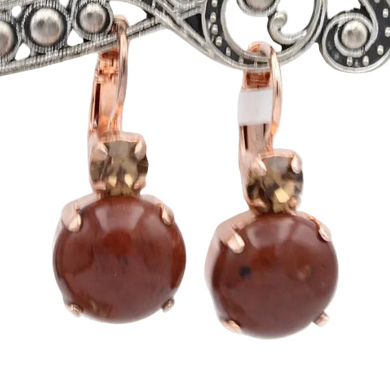 A pair of rose gold earrings with round brown stone drops, hanging from an ornate silver display stand against a white background.