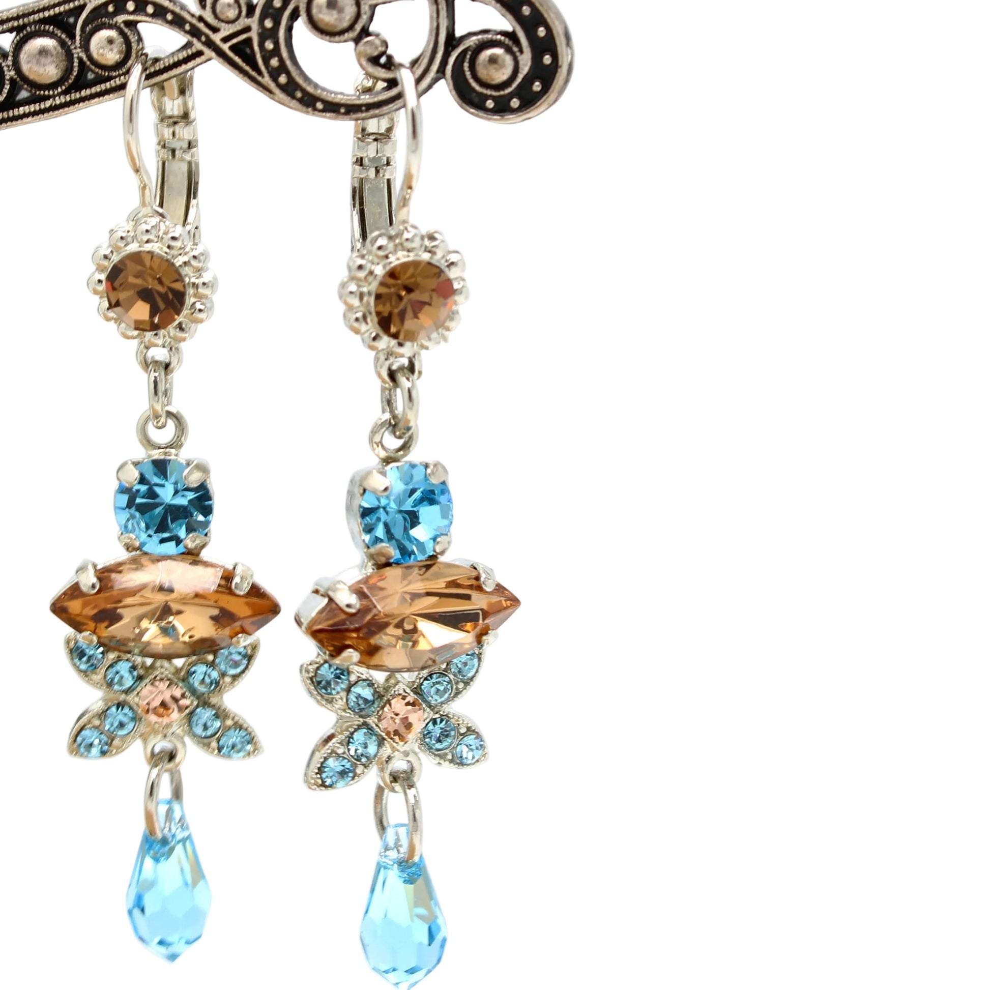 A pair of dangling earrings with ornate silver-toned metalwork, amber-brown marquise-cut crystals, light blue round crystals, and light blue teardrop dangling drops, shown hanging from a decorative display stand against a white background.