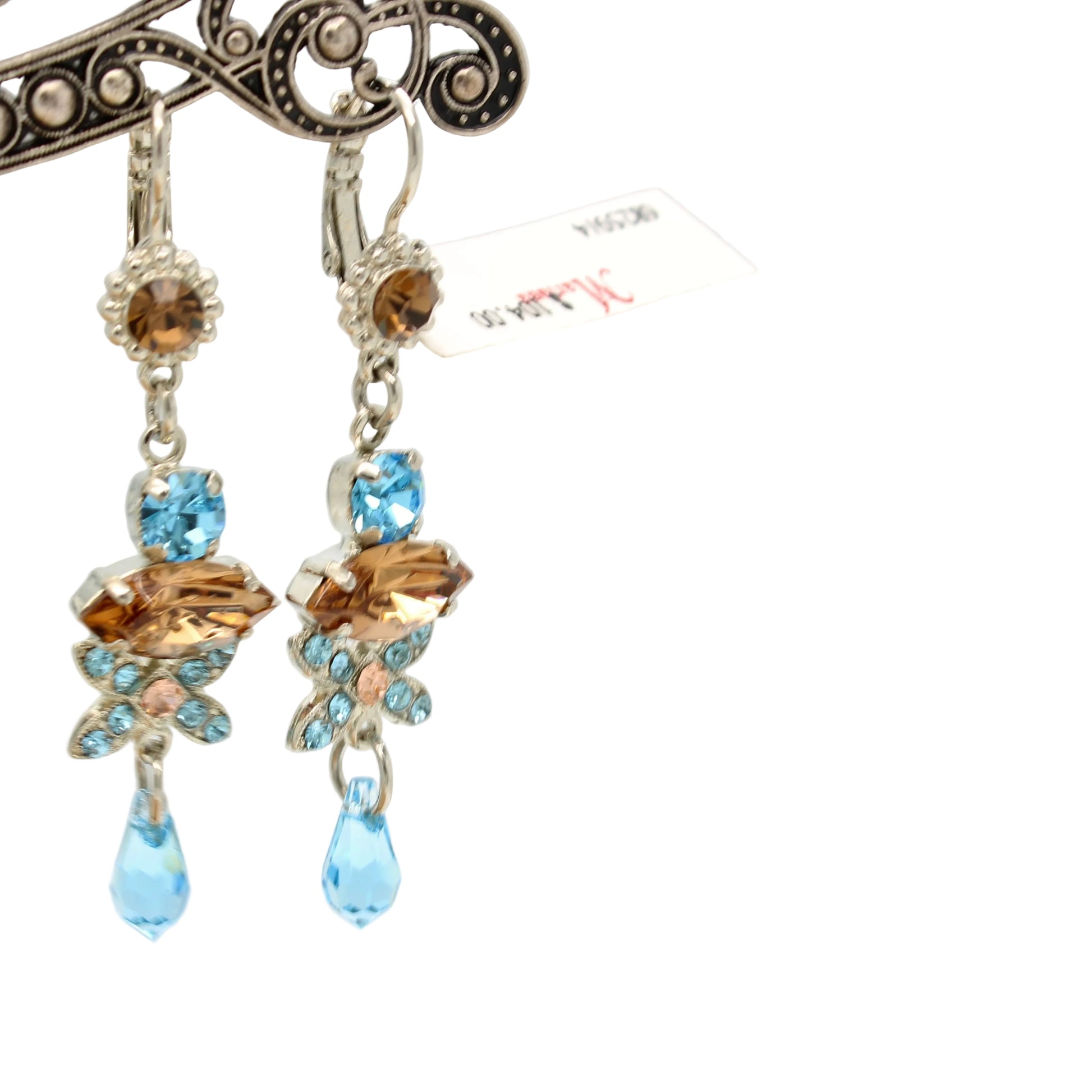 A pair of dangling earrings with silver-tone metal, light blue faceted gemstones, and amber-brown crystal flower motifs, hanging from an ornate display stand against a light gray background.