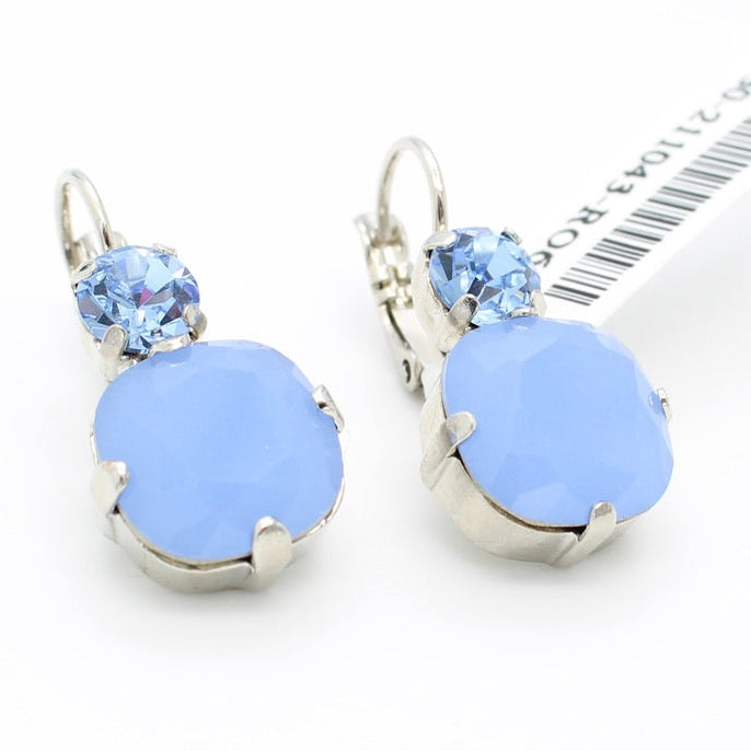 A pair of silver leverback earrings featuring a large light blue oval stone and a smaller round light blue crystal, displayed on a white background next to a barcode card.