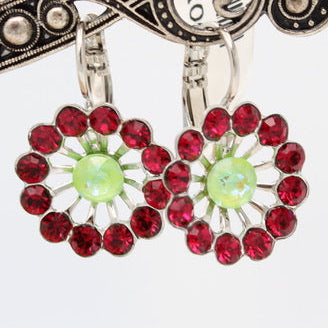 A pair of silver-toned dangle earrings with red faceted gemstones in a circular floral pattern and light green center stones, displayed on an ornate silver stand engraved with 'Mariana Spirit of Design'.