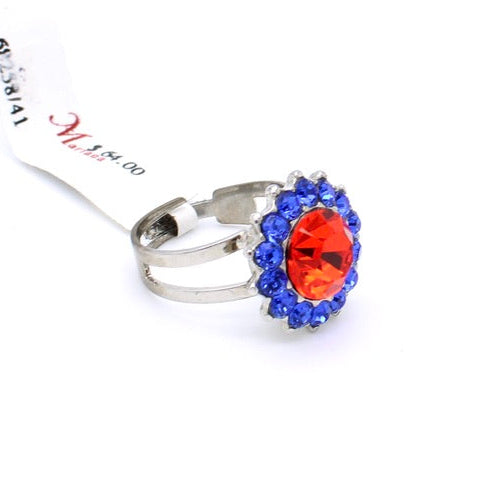 A silver-toned ring featuring a large oval orange-red center stone surrounded by a halo of small blue stones, with a white tag attached, on a plain white background.